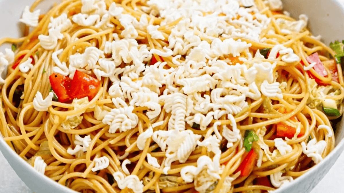 A bowl of stir-fried noodles with colorful vegetables, topped with crumbled uncooked instant ramen noodles.