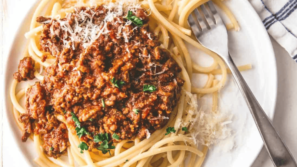 A plate of spaghetti topped with meat sauce and grated parmesan cheese, garnished with herbs. A fork rests on the plate beside the pasta.