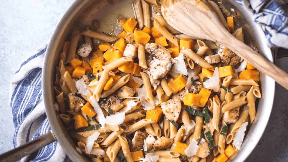 A skillet filled with penne pasta, chunks of chicken, diced butternut squash, spinach, and shaved Parmesan cheese, with a wooden spoon resting on the side.