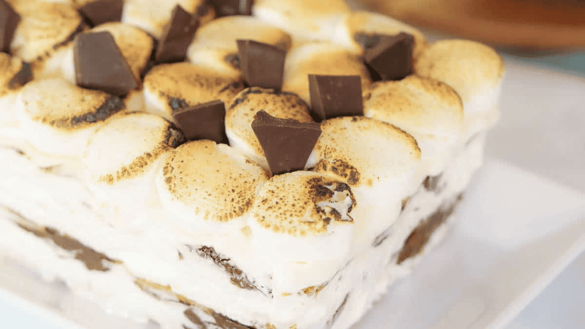 A close-up of a dessert topped with toasted marshmallows and chunks of chocolate, layered with creamy filling, resembling a s'mores icebox cake on a white plate.