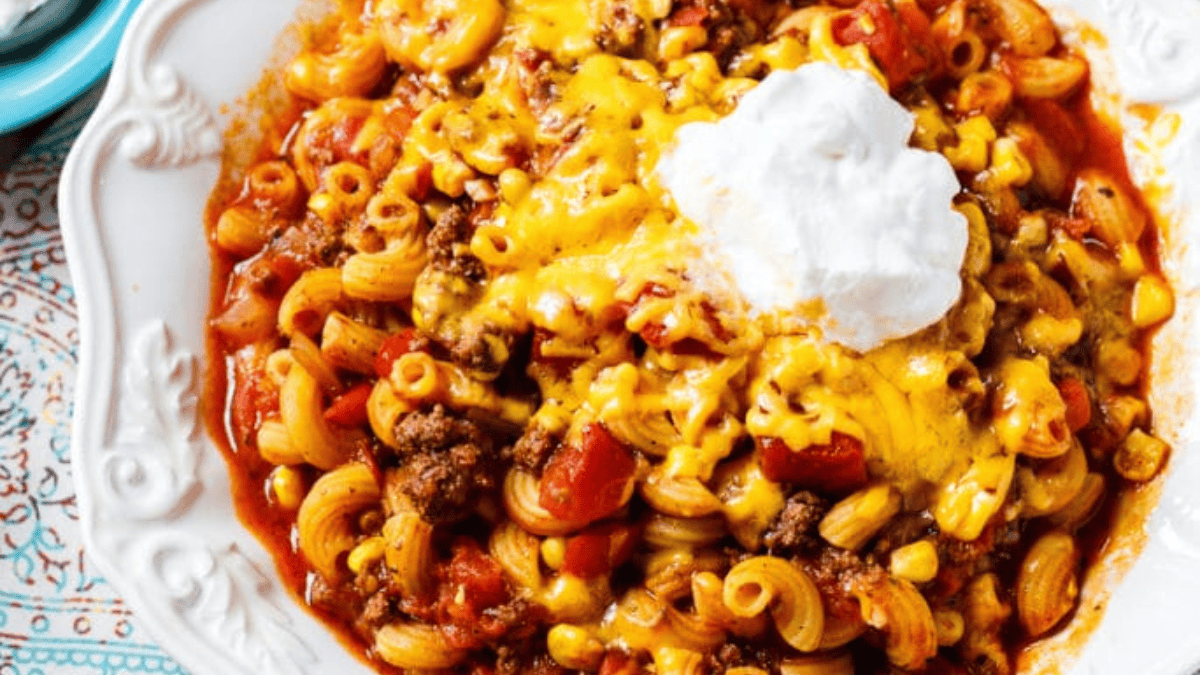 A plate of cheesy macaroni with ground beef, corn, diced tomatoes, and melted cheddar cheese, topped with a dollop of sour cream.