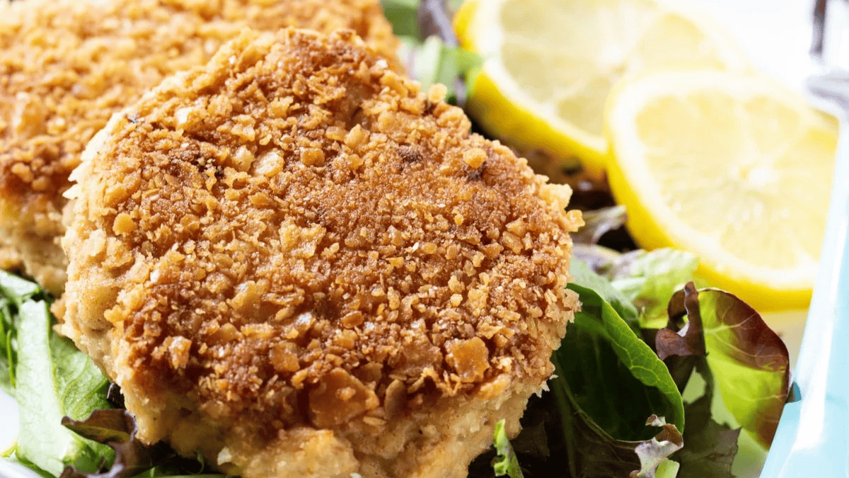 A close-up of two golden-brown, crispy crab cakes served on a bed of mixed greens, with slices of fresh lemon in the background.
