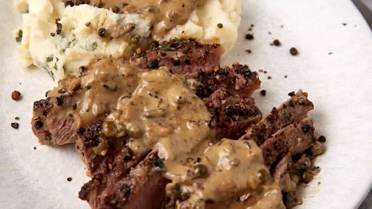 Sliced steak au poivre topped with peppercorn sauce served next to creamy mashed potatoes on a white plate, with a fork and a piece of bread visible in the background.