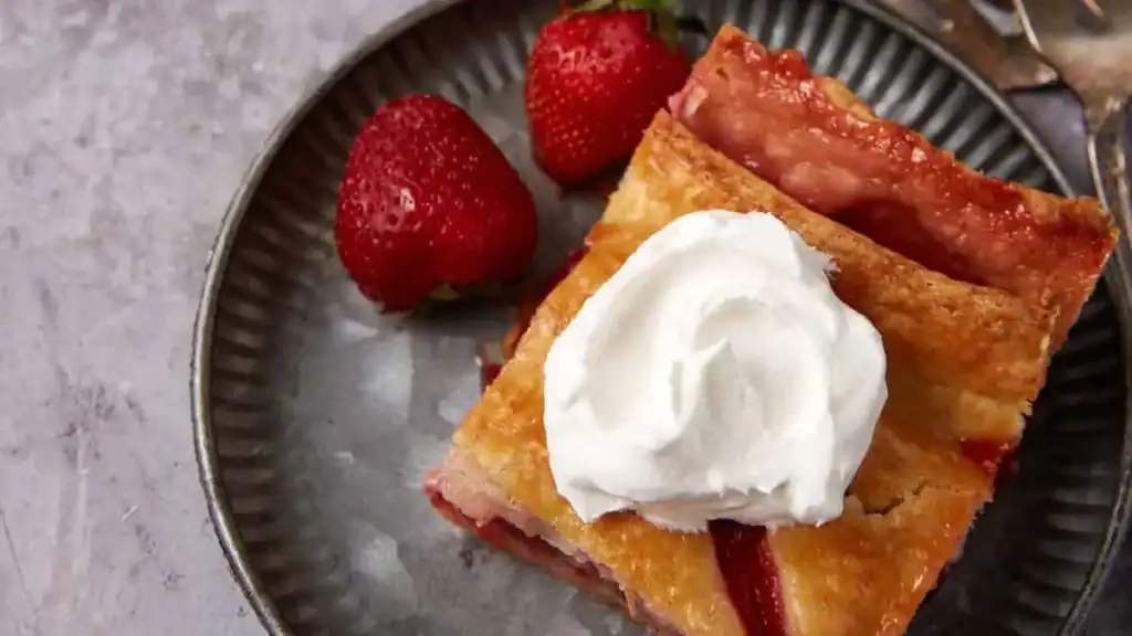 A slice of strawberry pie topped with whipped cream on a metal plate, with two fresh strawberries beside it.