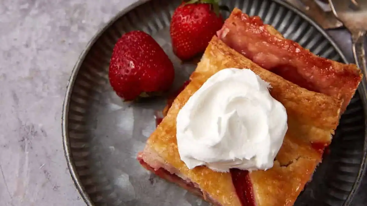 A slice of strawberry pie topped with whipped cream on a metal plate, with two fresh strawberries beside it.