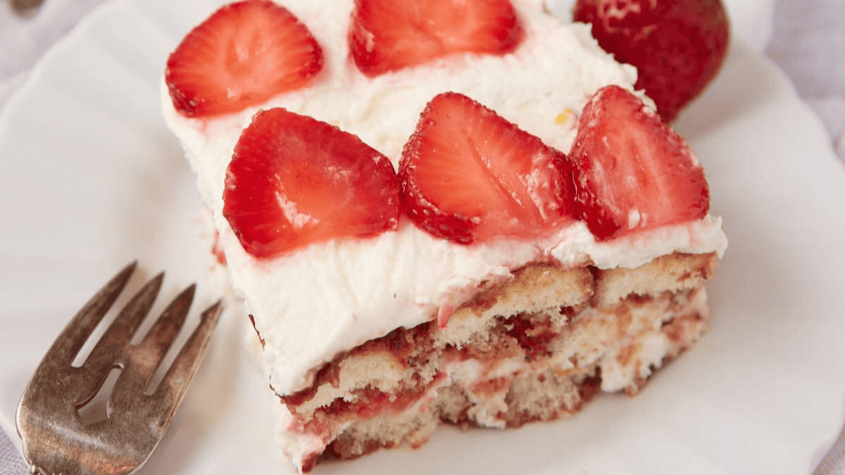 A slice of strawberry tiramisu on a white plate, topped with sliced strawberries and creamy layers, with a fork placed beside it. A whole strawberry is in the background.