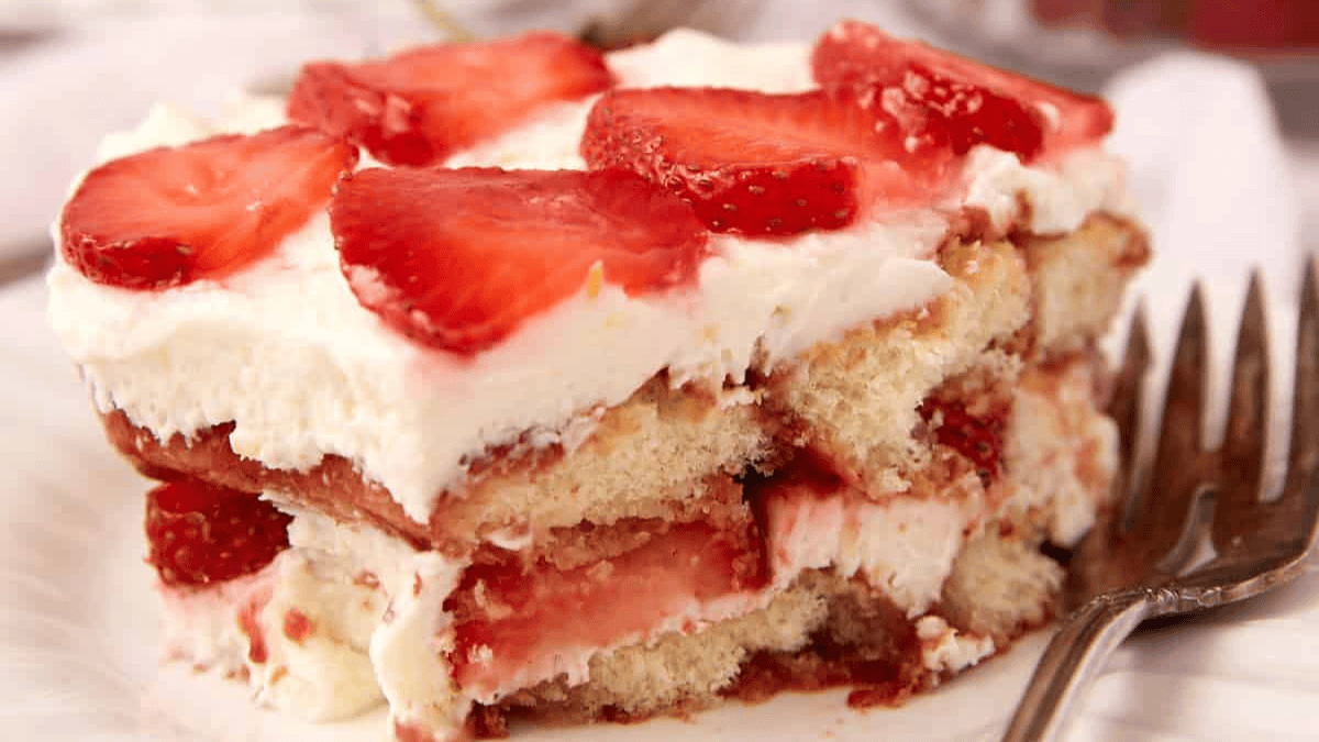 A close-up of a slice of strawberry tiramisu, showing layers of sponge cake, creamy filling, and fresh strawberry slices on top, with a fork beside it on a white plate.