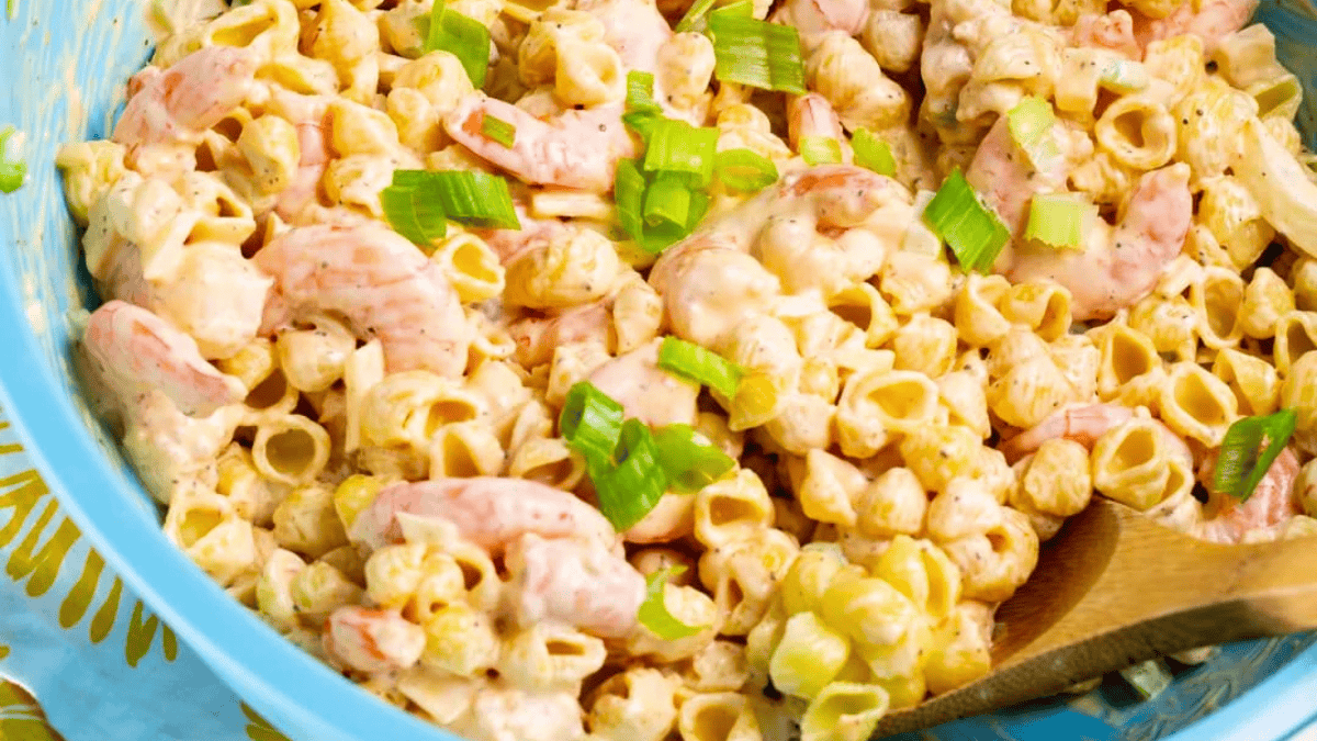 A close-up of creamy pasta salad with shrimp, small shell pasta, chopped green onions, and a wooden spoon in a blue bowl.