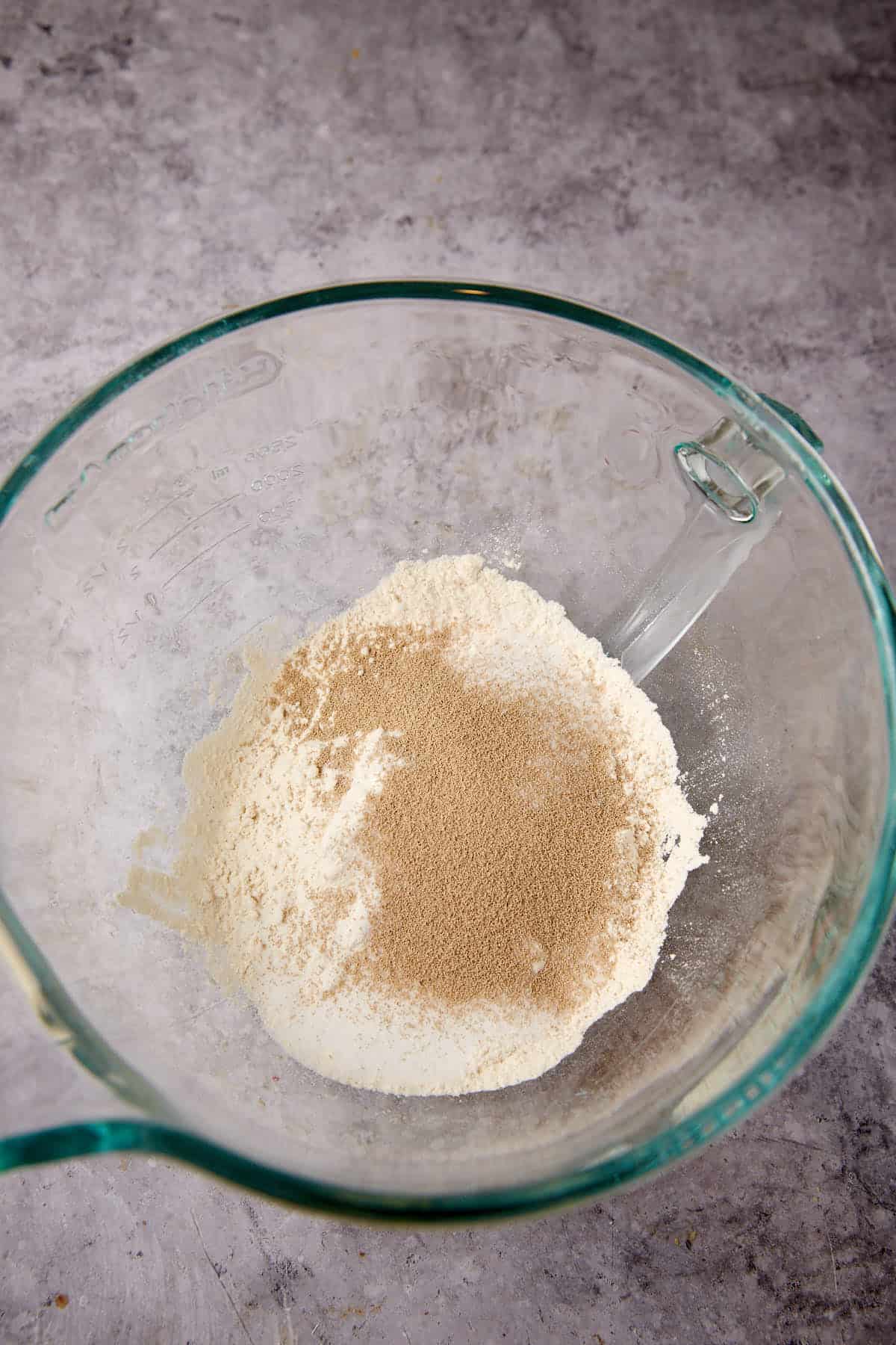 A glass mixing bowl containing a mound of flour with dry yeast sprinkled on top, placed on a gray countertop.