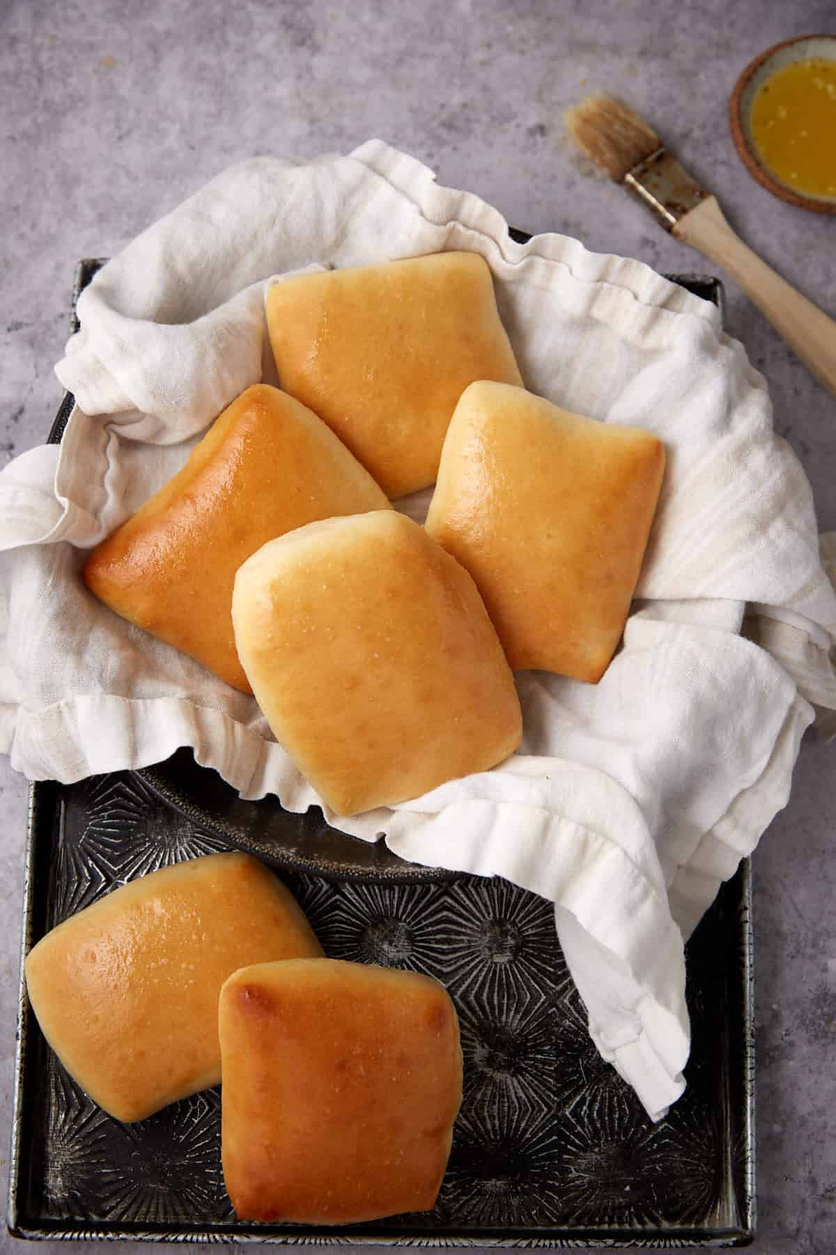 A tray lined with a white cloth holds six golden-brown, square dinner rolls. A small bowl of melted butter and a pastry brush are placed nearby on a light grey surface.