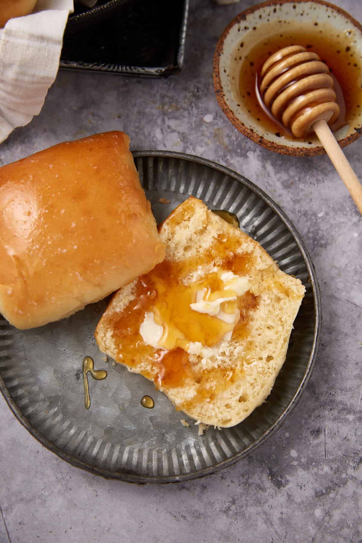 A metal plate holds a soft dinner roll and another half spread with butter and drizzled with honey. A wooden honey dipper rests in a small bowl filled with honey beside the plate.