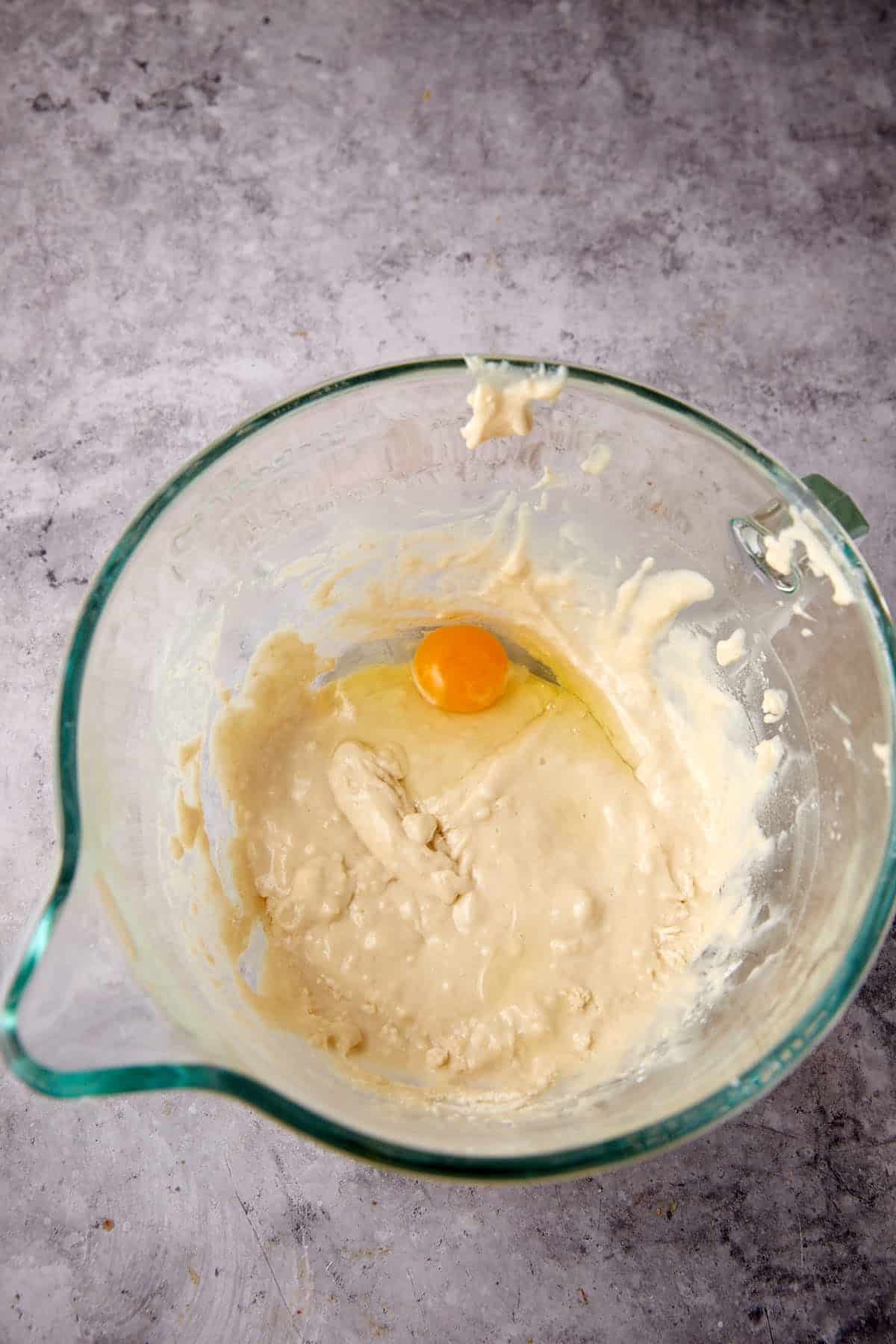 A glass mixing bowl containing pale, thick batter with a whole raw egg on top, ready to be transformed into delicious Texas Roadhouse Rolls, set on a gray countertop.