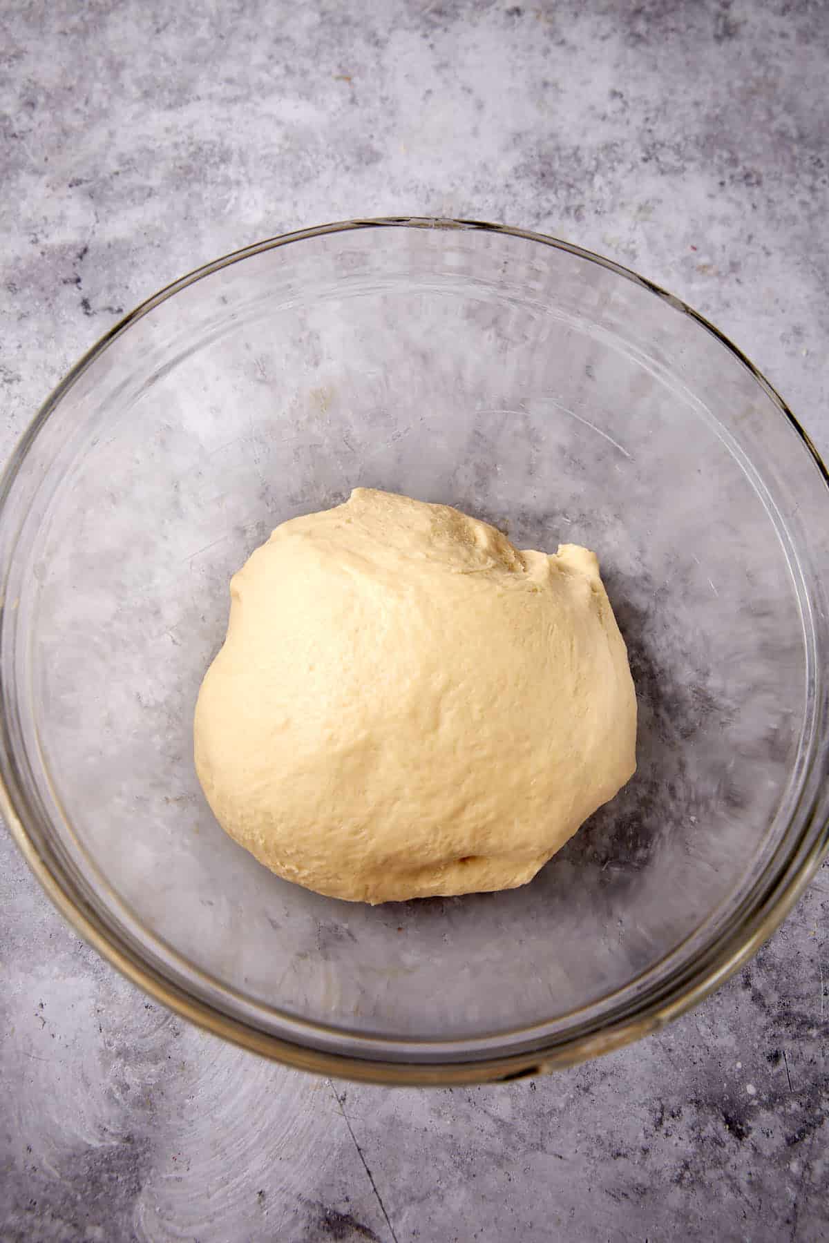 A ball of smooth dough rests in a large, clear glass bowl set on a mottled gray countertop.