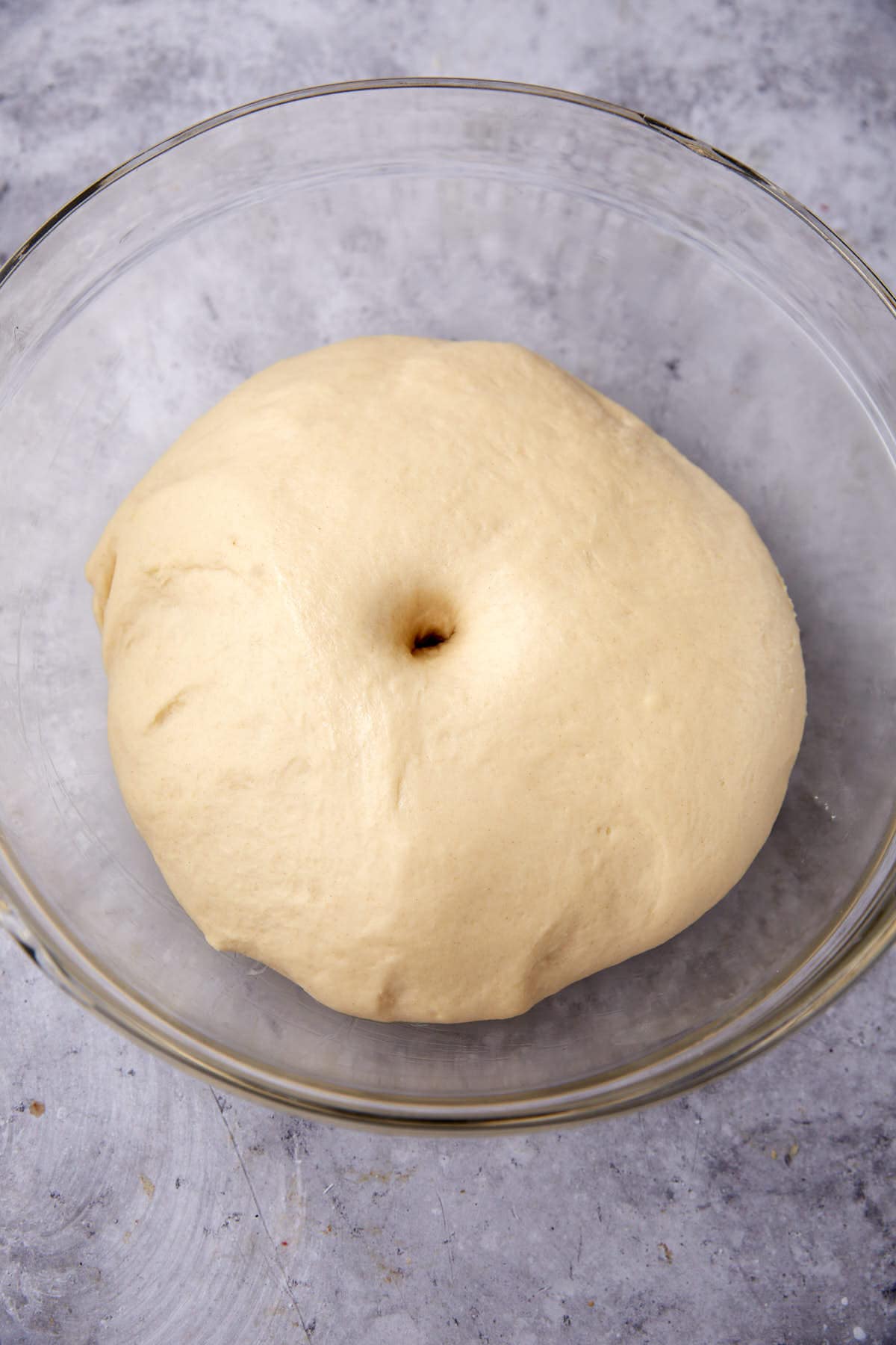 A ball of risen dough with a finger indentation in the center sits in a clear glass bowl on a lightly textured gray surface.