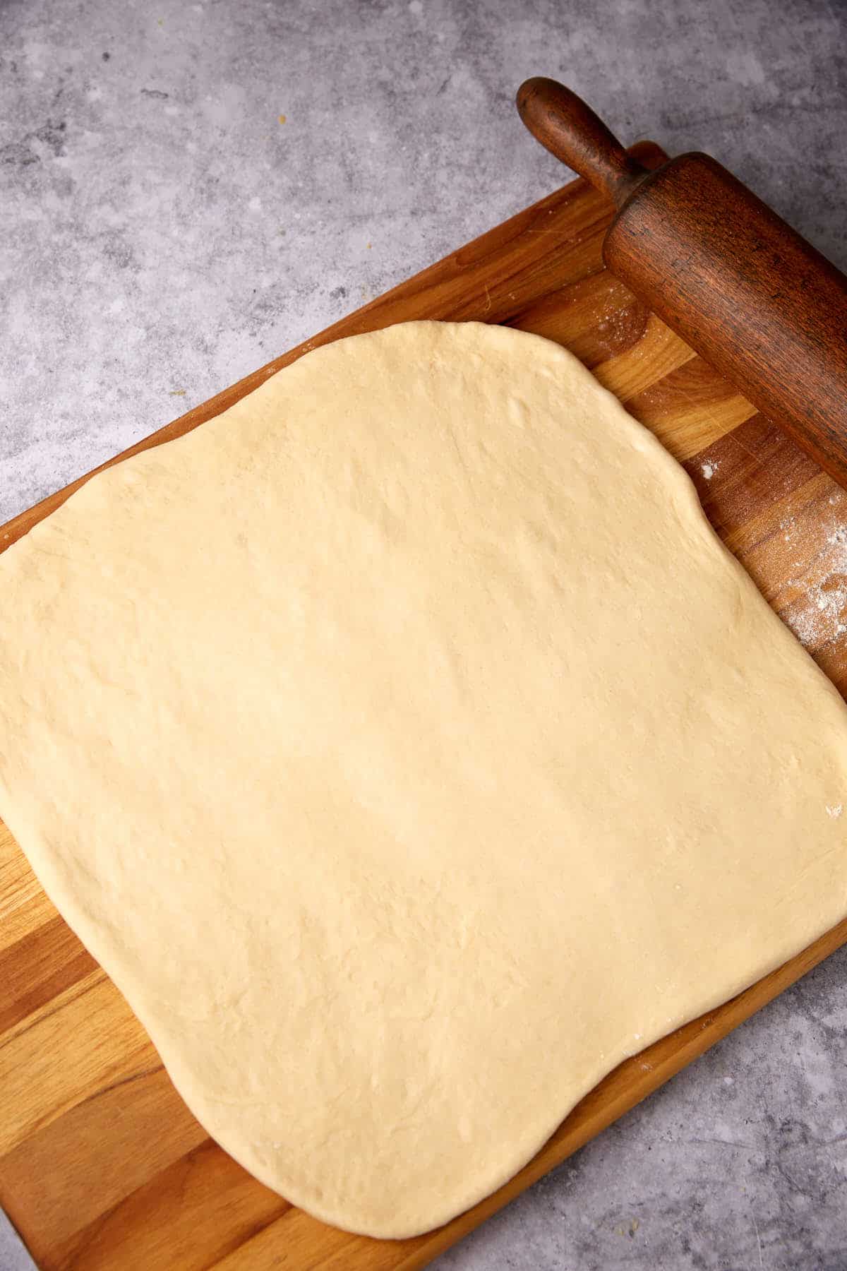 A sheet of dough rolled out into a large rectangular shape on a wooden cutting board, with a wooden rolling pin resting at the top right corner.