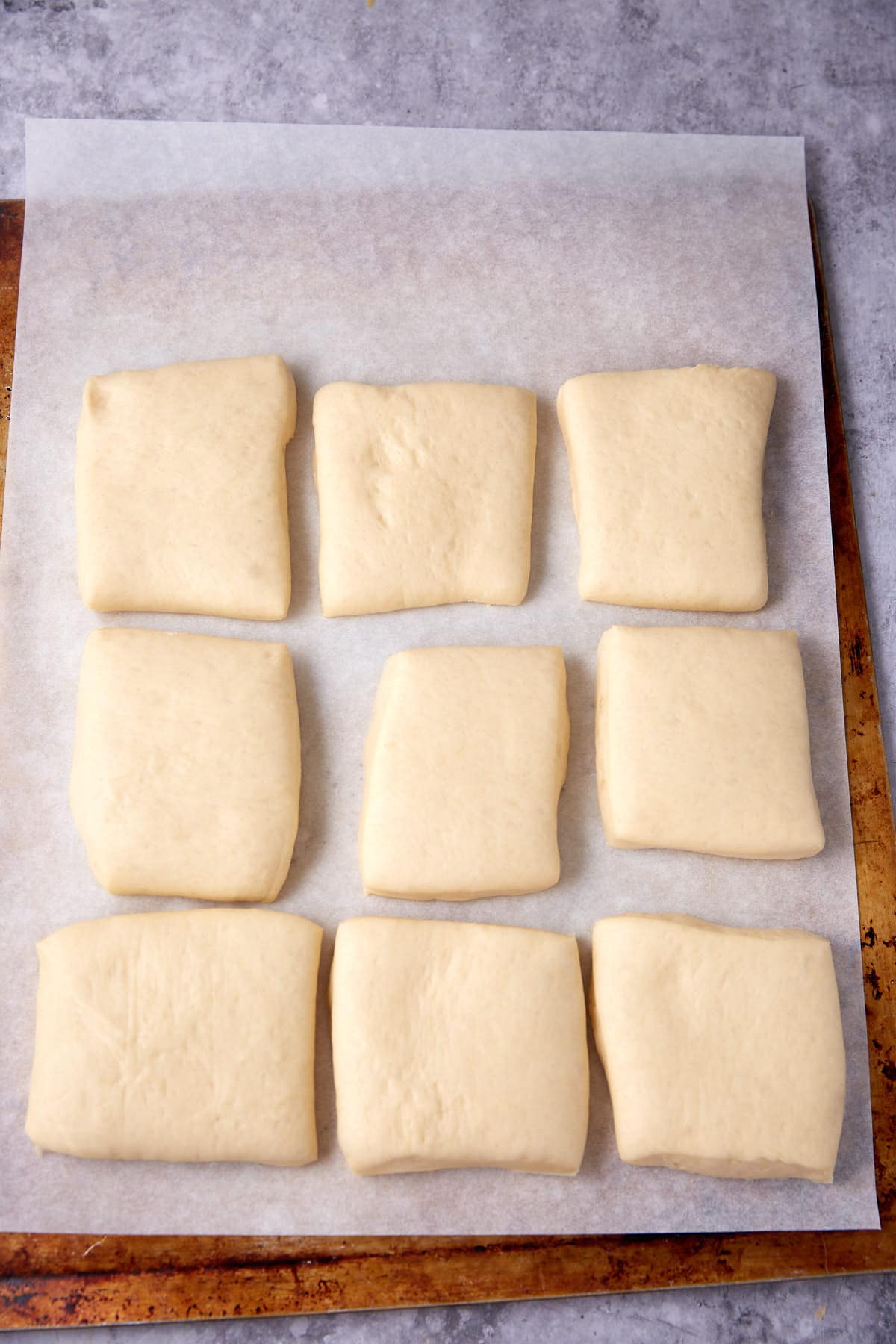 Nine evenly spaced, square pieces of raw dough sit on a sheet of parchment paper atop a baking tray, ready to be baked.