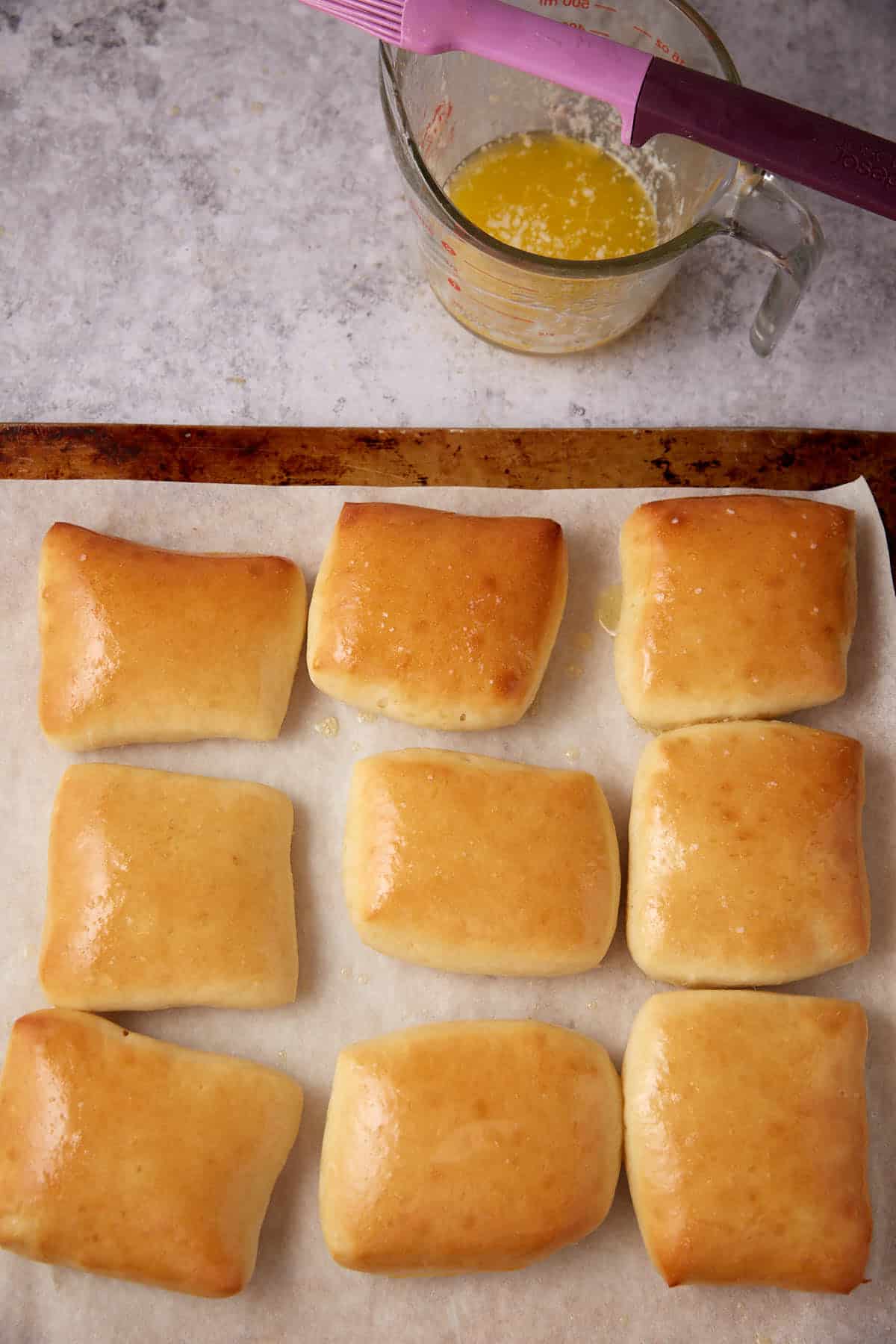 Nine golden-brown rectangular rolls are arranged on parchment paper atop a baking sheet. A cup of melted butter with a pink brush rests nearby on a gray surface.