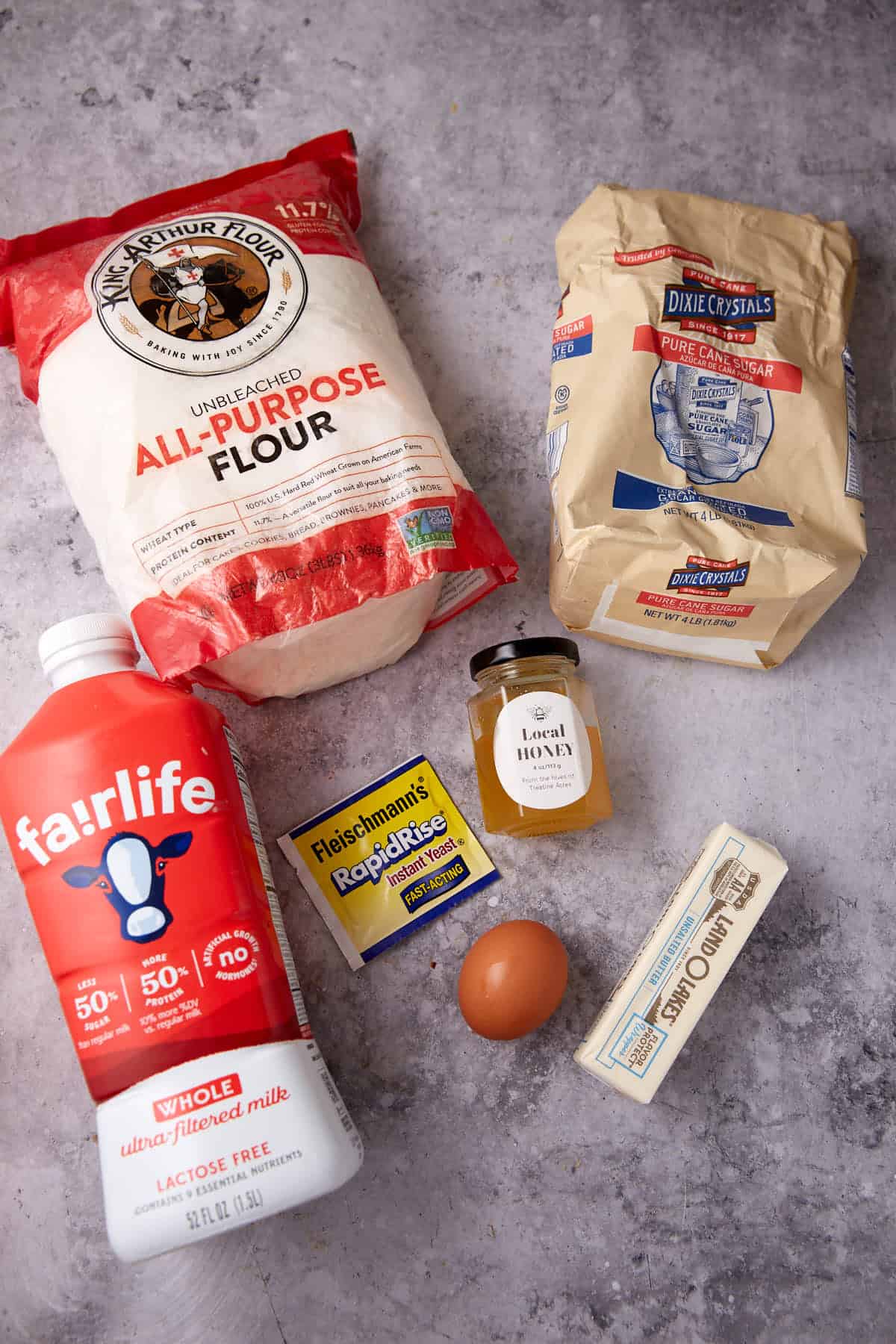 Baking ingredients on a gray surface: a bag of all-purpose flour, a bag of sugar, a bottle of milk, a jar of honey, a packet of yeast, a packet of rapid rise yeast, a stick of butter, and a brown egg.