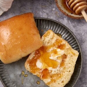 A buttered Texas Roadhouse Roll split open on a metal plate, with melted butter and honey drizzled on top. Another whole roll sits beside it, while a honey dipper with honey rests in the background on the gray surface.