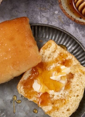 A buttered Texas Roadhouse Roll split open on a metal plate, with melted butter and honey drizzled on top. Another whole roll sits beside it, while a honey dipper with honey rests in the background on the gray surface.
