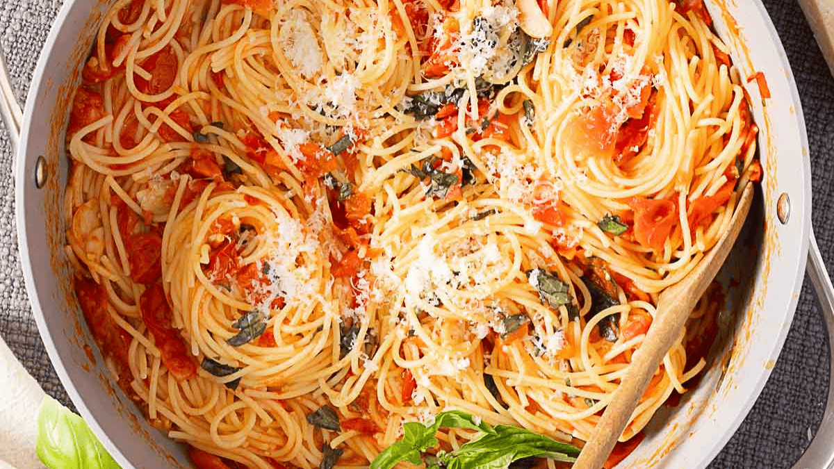 A skillet filled with cooked spaghetti in tomato sauce, garnished with grated cheese and fresh basil, with a wooden spoon resting inside.