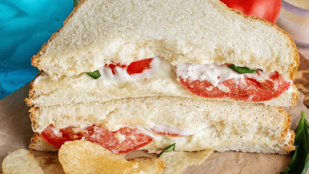 A close-up of a tomato sandwich with mayonnaise and fresh basil on white bread, cut in half and stacked, with potato chips beside it on brown parchment paper.
