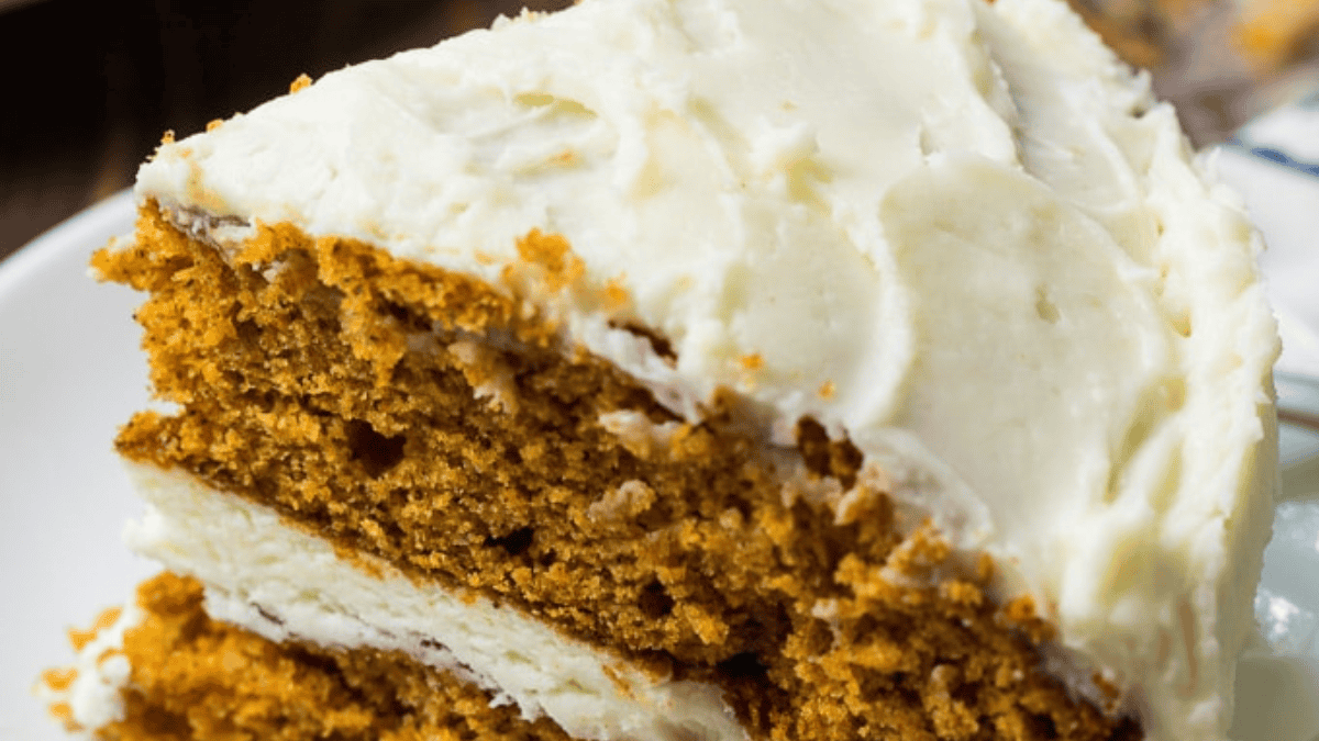 A close-up of a slice of two-layer carrot cake with creamy white frosting between the layers and on top, served on a white plate.