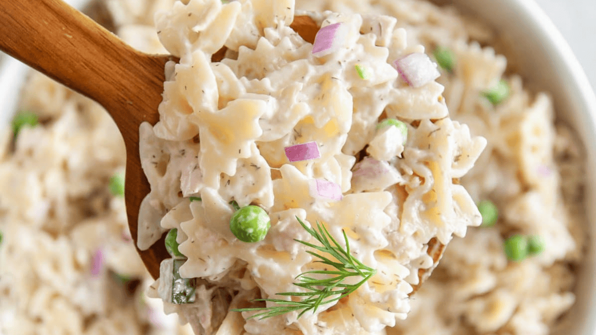 A close-up of a wooden spoon holding creamy bowtie pasta salad with green peas, red onion pieces, and a sprig of fresh dill. The background shows a bowl filled with more of the pasta salad.