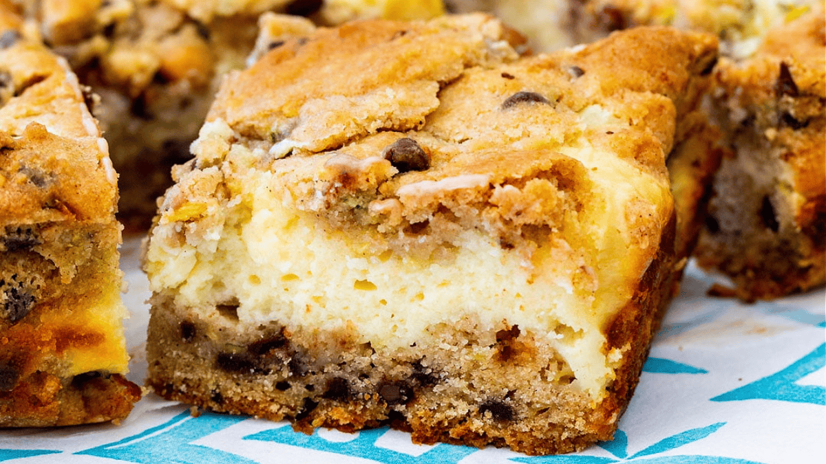 A close-up of a thick, square dessert bar with a crumbly top, creamy middle layer, and a bottom layer containing chocolate chips, resting on a blue and white patterned surface.