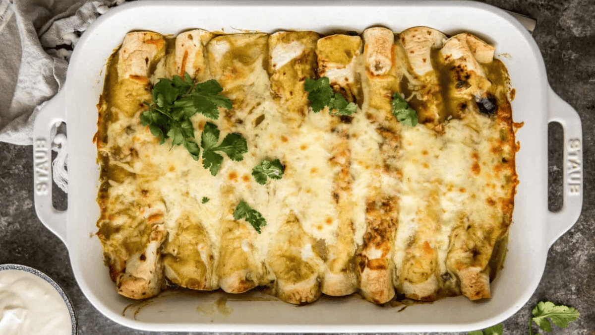 A baking dish filled with enchiladas topped with melted cheese and garnished with fresh cilantro, with a green sauce visible on top and to the sides.