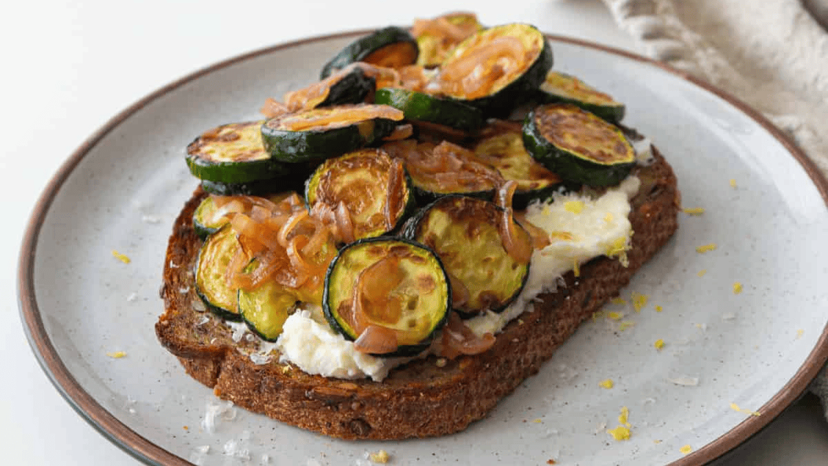 A slice of whole grain toast topped with creamy ricotta cheese, sautéed zucchini rounds, and caramelized onions, served on a white plate with a brown rim.
