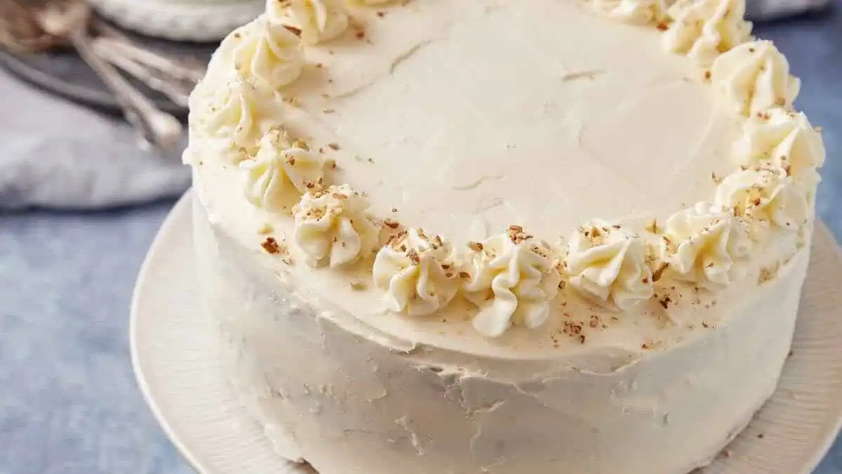 A round frosted cake decorated with swirls of white icing and a sprinkle of crumbs on top, displayed on a white plate against a light blue background.
