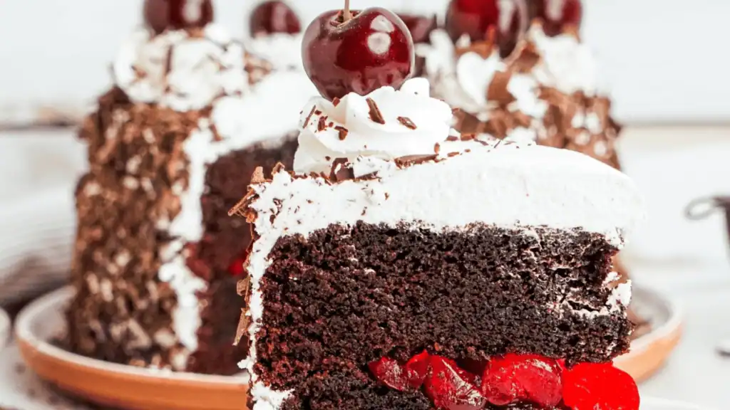 A close-up of a slice of Black Forest cake with layers of chocolate sponge, whipped cream, cherries, and chocolate shavings, topped with a cherry. The whole cake is visible in the background.