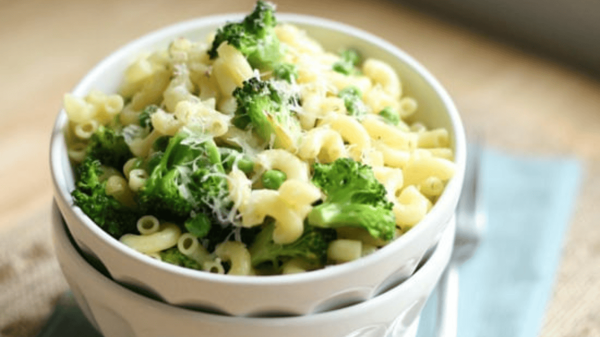 A white bowl filled with macaroni pasta, broccoli florets, and green peas, topped with grated cheese, sits on a light blue napkin with a fork nearby.