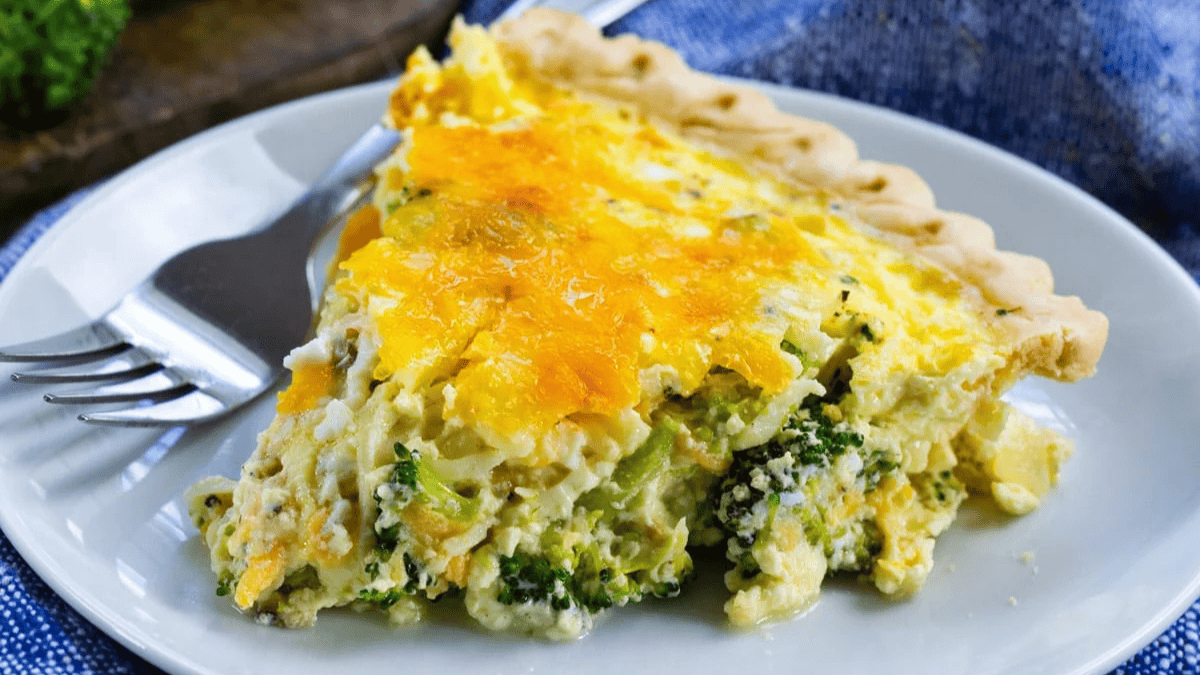 A slice of broccoli and cheese quiche sits on a white plate with a fork and knife beside it, showing a golden, cheesy top and a flaky crust.