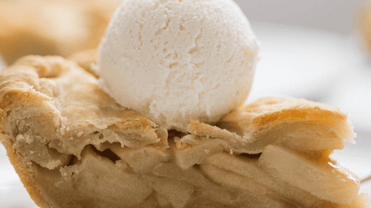 A close-up of a slice of apple pie topped with a scoop of vanilla ice cream, showing flaky crust and layered apple filling.