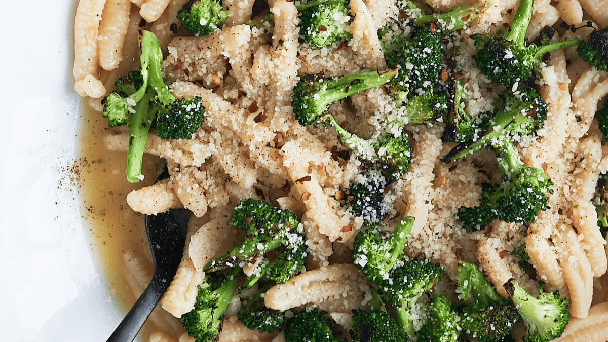 A plate of pasta topped with sautéed broccoli florets, grated cheese, red pepper flakes, and cracked black pepper, with a black spoon resting on the side.
