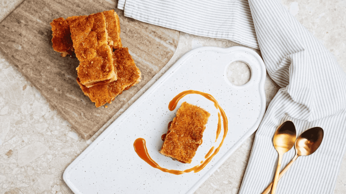 A rectangular caramel dessert bar is plated on a white cutting board with caramel sauce drizzled around it, next to a stack of bars on a marble slab, with two spoons and a striped cloth nearby.