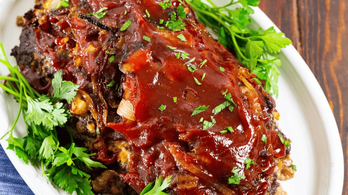 A glazed meatloaf topped with chopped parsley sits on a white platter, surrounded by fresh parsley sprigs. The meatloaf is covered in a shiny tomato-based sauce.