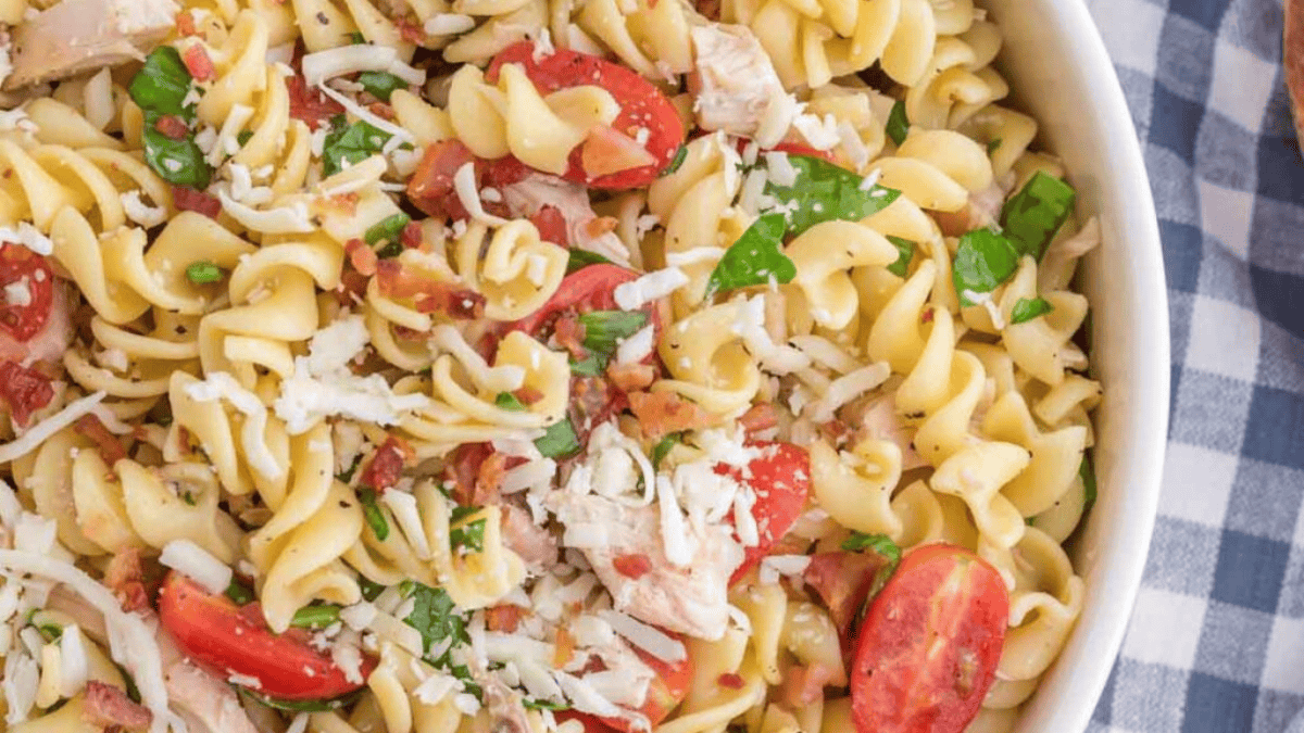 A close-up of a bowl of rotini pasta salad with cherry tomatoes, shredded cheese, spinach, bacon bits, and chicken pieces, on a checkered tablecloth.