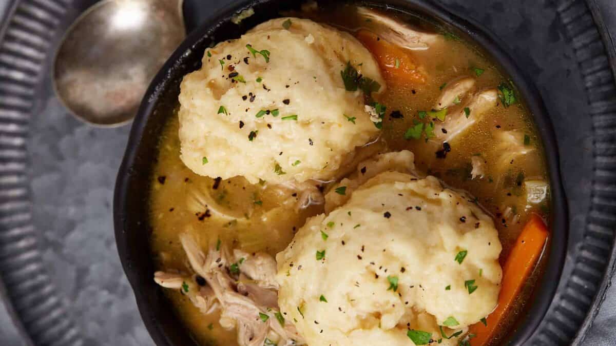 A bowl of hearty chicken and dumplings soup is shown. The bowl contains shredded chicken, vegetables like carrots, and two large, fluffy dumplings. The dish is garnished with chopped parsley and fresh ground black pepper, and a spoon rests on the side.