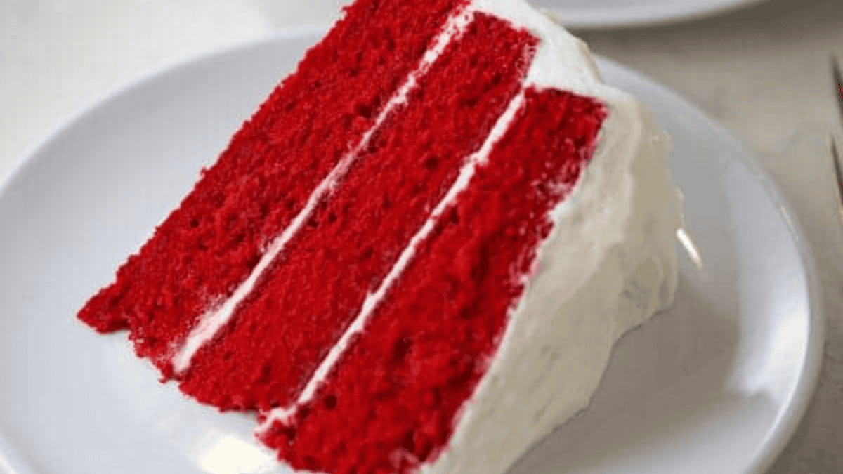 A slice of three-layer red velvet cake with white cream cheese frosting between the layers and on the outside, served on a white plate.