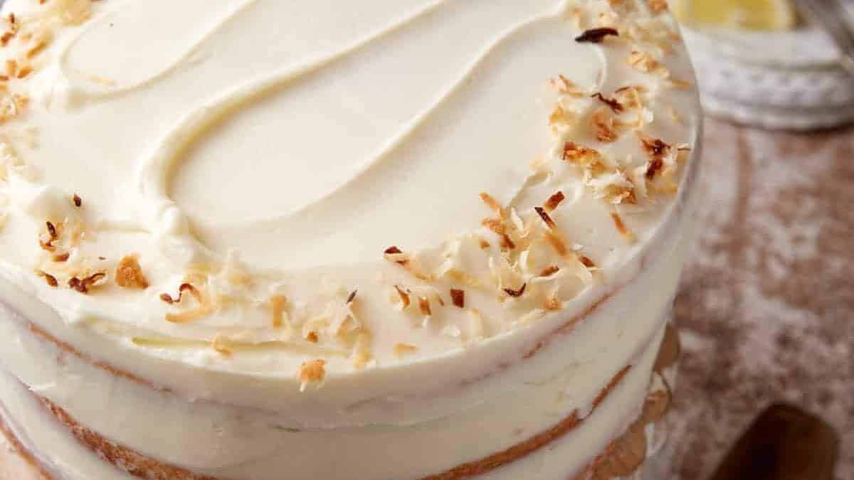 A close-up of a frosted layer cake topped with toasted coconut flakes, showing creamy white icing and partially visible cake layers.