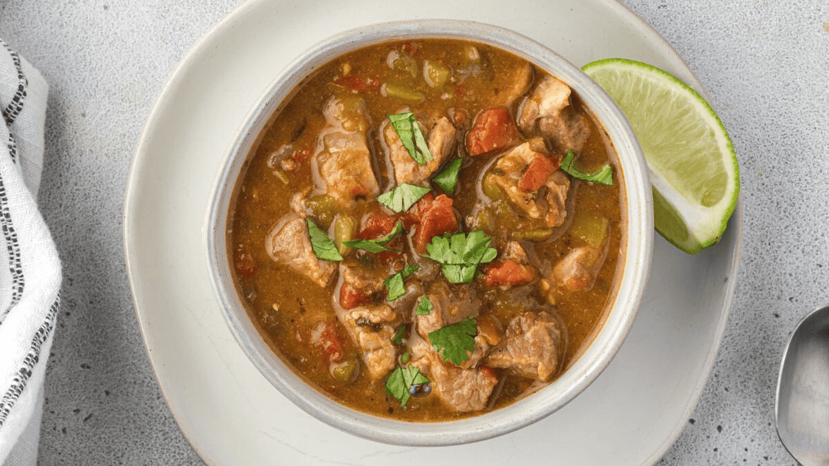 A bowl of stew with chunks of meat, tomatoes, green peppers, and herbs, garnished with fresh cilantro, sits on a white plate with a wedge of lime on the side. A spoon and napkin are nearby.