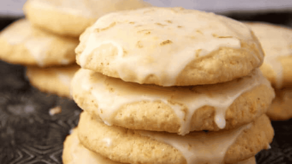 A close-up of a stack of soft, round cookies topped with a thin layer of white glaze, with more cookies blurred in the background.