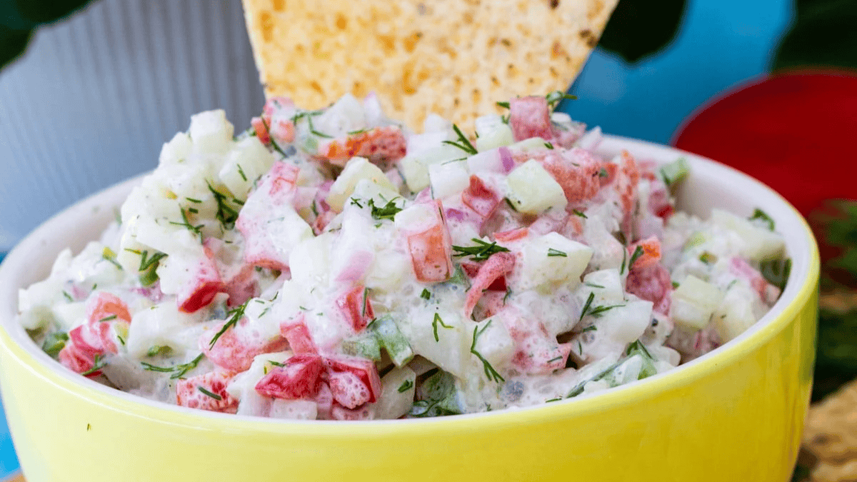 A bowl of creamy vegetable dip with chopped cucumbers, red peppers, and herbs, topped with a tortilla chip, served in a yellow dish.