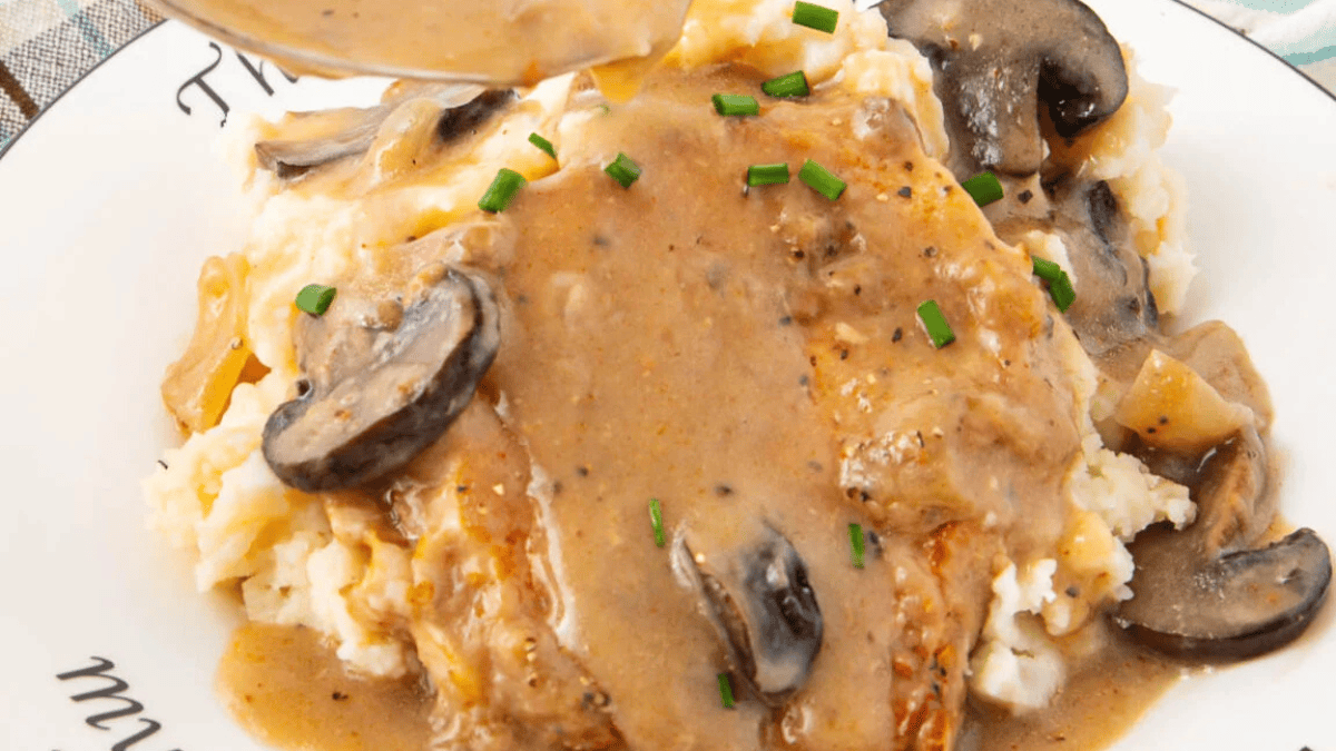 A close-up of mashed potatoes topped with a creamy mushroom gravy, sliced mushrooms, and chopped chives on a white plate.