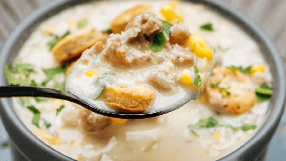 A close-up of a spoonful of creamy soup with corn, ground meat, herbs, and round crackers, held above a bowl filled with the same soup and garnished with fresh parsley.
