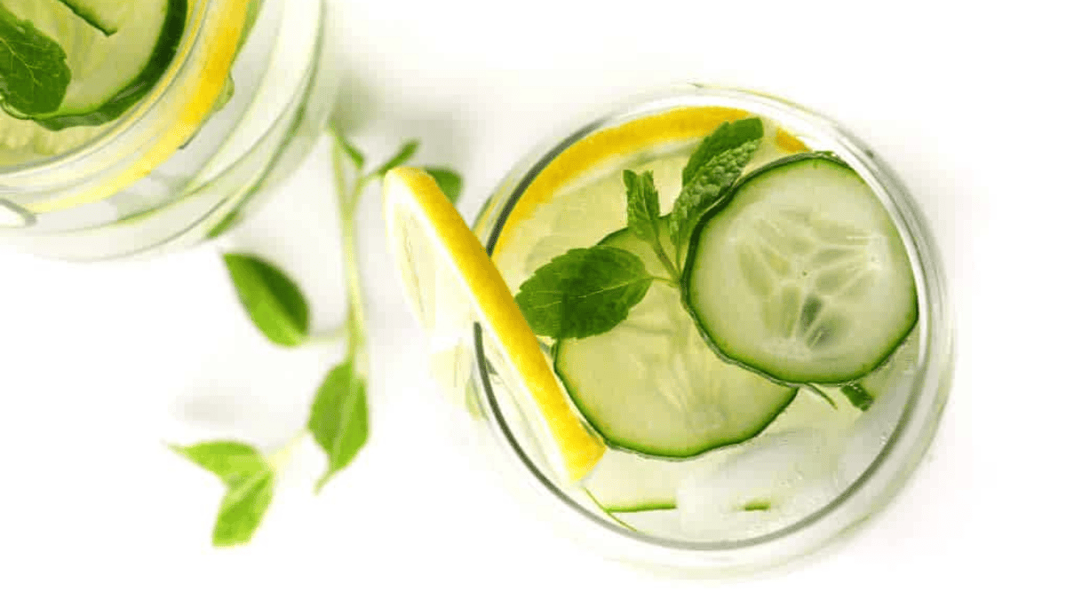 A glass of water with ice, cucumber slices, lemon slices, and fresh mint leaves on a white surface. Another similar glass and extra mint leaves are partially visible beside it.