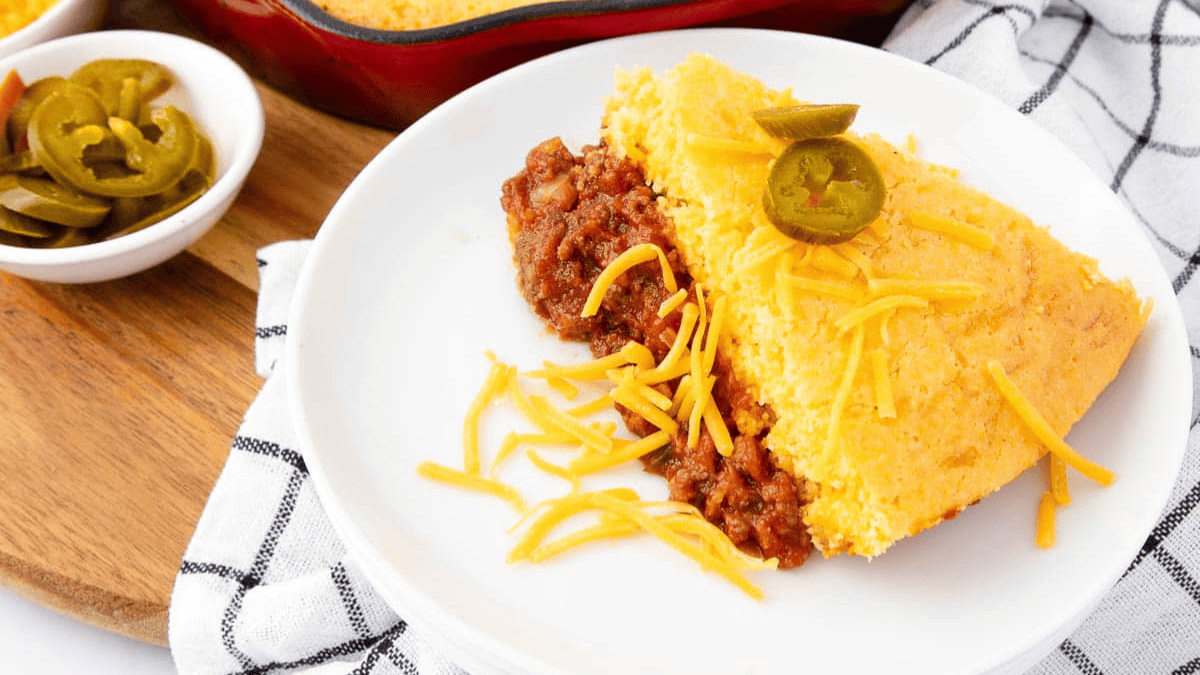 A slice of cornbread casserole topped with shredded cheddar cheese and jalapeno slices sits on a white plate; a bowl of jalapenos and a casserole dish are nearby on a checkered cloth.